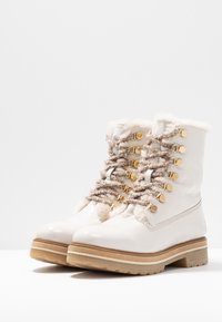 Botas de charol blanco con ribete de piel de piel de oveja, cordones beige y herrajes dorados. Suela gruesa y texturizada con un diseño de banda de rodadura robusto.