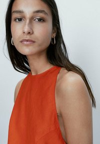 Pomarańczowy bezrękawnik o gładkiej fakturze i wysokim dekolcie; modelka nosi małe, złote kolczyki w kształcie kółek; neutralne tło.