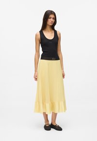 Débardeur noir avec encolure dégagée, assorti à une jupe jaune plissée avec un ourlet en satin. Le mannequin porte des ballerines en maille noire.