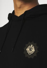 Black hoodie with a drawstring, featuring a gold graphic design of an eye and rays, on a soft, textured fabric.
