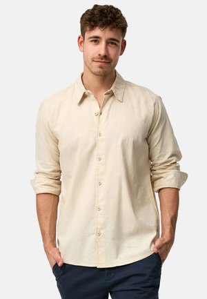 Homme aux cheveux courts et bouclés portant une chemise beige à boutons avec les manches retroussées jusqu’aux coudes et un pantalon bleu marine, les mains dans les poches.