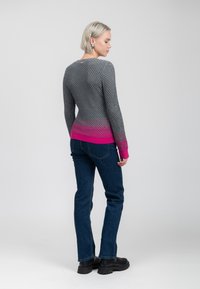 Vrouw met kort blond haar, gekleed in een getextureerde grijze trui met roze manchetten en zoom, donkerblauwe jeans en zwarte laarzen, met de rug naar ons toegekeer.