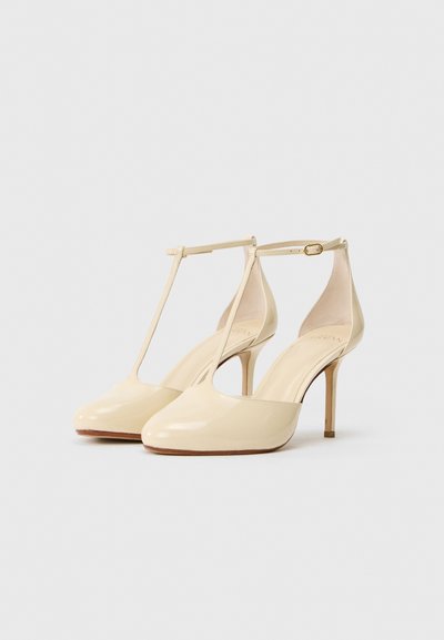 Beige patent leather high heels with a T-strap design, pointed toe, and slender heel. Smooth texture with minimal hardware accents.