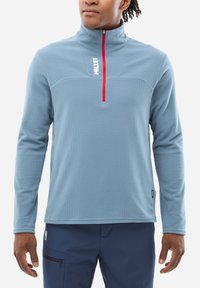 Pull en polaire bleu clair avec un col zippé, tissu texturé, accents de fermeture éclair rouges et un logo blanc sur la poitrine. Manches longues.