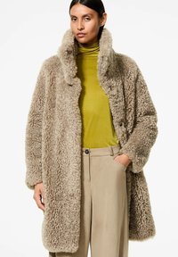 Cappotto in finta pelliccia color marrone con colletto alto, chiusura a bottoni e tasche laterali, indossato sopra un dolcevita verde e pantaloni beige.