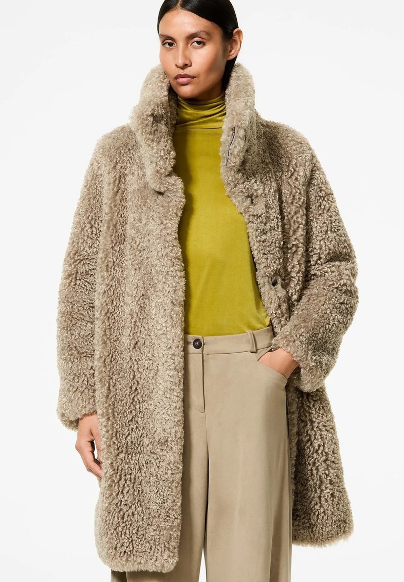 Cappotto in finta pelliccia color marrone con colletto alto, chiusura a bottoni e tasche laterali, indossato sopra un dolcevita verde e pantaloni beige.