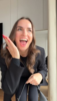 Svart blazer over en svart topp, svarte bukser, holder en svart veske med gullakenter. Langt, bølgete brunt hår og klare røde negler. Tresparkel.