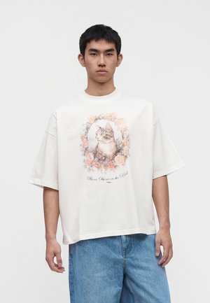 Jeune homme portant un T-shirt blanc oversize avec un portrait de chat encadré de roses roses et un jean bleu, debout devant un fond uni.