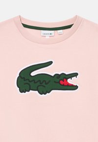 Sudadera de color rosa claro con un logo de cocodrilo verde y acentos rojos, textura suave y un cuello redondo acanalado con ribete verde.
