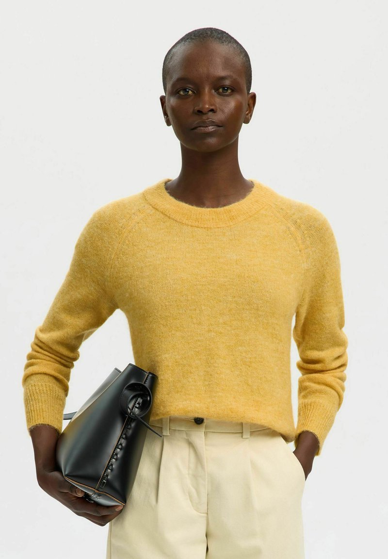 Personne portant un pull jaune et un pantalon beige tenant un sac à main noir de la main droite, sur un fond blanc uni.