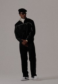 Homme portant un béret noir, des lunettes, un survêtement noir avec des liserés blancs, un col roulé blanc, des baskets blanches avec un swoosh noir, se tenant devant un fond uni.