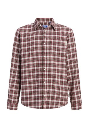 Ung person med krøllet hår, der bærer en rød ternet skjorte over en brun t-shirt, sorte bukser og tanfarvede støvler, står og kigger til siden.