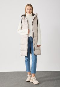 Puffervest i lys beige med hette, med glidelås og vertikal søm. Bært over en hvit turtleneck og blå jeans.