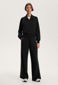 Fekete tréningruha puhány anyagból, fehér díszítéssel ellátott, cipzáros dzsekivel és laza szabású nadrággal, amely rugalmas derékkal rendelkezik.