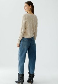 Beige gestrickter Pullover mit strukturiertem, rautenförmigem Design und Fransen-Details, kombiniert mit blauen hoch taillierten Jeans und schwarzen Knöchelstiefeln.
