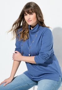 Modrý rolák s dlouhým rukávem z texturované tkaniny, s zatočenými rukávy a zakřiveným lemem, doplněný světle modrými džínami a stříbrným náhrdelníkem.