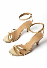 Paar goldfarbene, metallische, offene High-Heel-Sandalen mit dünnen Knöchelriemen und mittelhohen Blockabsätzen auf weißem Hintergrund.