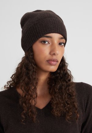 Jeune femme avec de longs cheveux bruns bouclés portant un bonnet en tricot marron foncé et un pull V côtelé assorti, sur un fond neutre.
