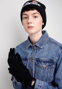 Černé pletené rukavice a čepice s logem "ellesse"; model má na sobě džínovou bundu. Zaměření na texturu a kontrastní barvy.