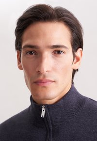 Giovane con capelli scuri che indossa un maglione zip grigio scuro, guarda direttamente verso la fotocamera su uno sfondo chiaro e semplice.