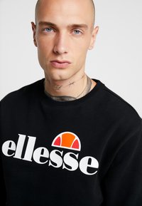 Schwarzes Sweatshirt mit weißem "ellesse"-Logo, das oben über dem Text einen stilisierten orange-weißen Akzent aufweist. Glatte Textur und Rundhalsausschnitt-Design.