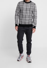 Schwarz-weißer karierten Strickpullover mit rundem Ausschnitt, gerippten Bündchen und schwarzem Saum, kombiniert mit dunklen Hosen und grauen Sneakers.