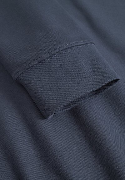 Poignet de chemise à manches longues bleu marine avec une texture lisse, présentant des coutures nettes et une coupe ajustée. Le matériau semble doux et extensible.