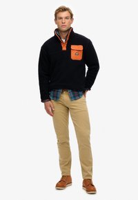 Marineblauer Fleece-Pullover mit einer orangen Nylon-Brusttasche, kombiniert mit einem blau karierten Hemd und beigen Hosen, abgerundet durch braune Schnürschuhe.