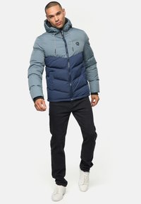Puffjakke med en teal øverste del og navy nederste del, med lynlås, hætte og quiltet design. Bæres med sorte bukser og hvide sko.