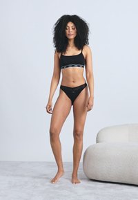 Crni sportski grudnjak sa širokim pojasom, uparен s crnim bikini gaćicama koje imaju vezicu. Glatka tekstura, uski kroj, minimalni detalji.