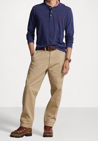 Mörkblå långärmad henley-tröja, beige cargobyxor, brun läderbälte och robusta bruna stövlar med röda snören. Har en tryckt krage som accent.