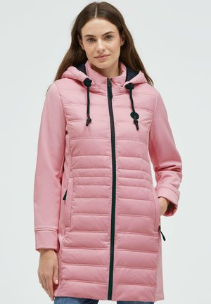 Frau trägt eine lange pinke gesteppte Jacke mit Kapuze, schwarzem Reißverschluss und Kordelzügen, Hände in den Taschen, neutraler Hintergrund.