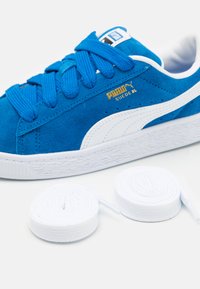 Kék velúrbőr sneaker fehér részletekkel, texturált talppal és vastag kék fűzőkkel. Két fehér cipőfűző is található a cipő mellett.