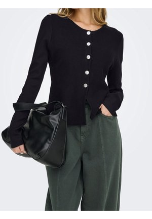 Femme portant un cardigan noir boutonné et un pantalon vert, tenant un grand sac en cuir noir avec une main dans la poche.