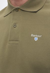Gros plan sur un polo Barbour vert olive avec deux boutons et logo de la marque brodé sur la poitrine.