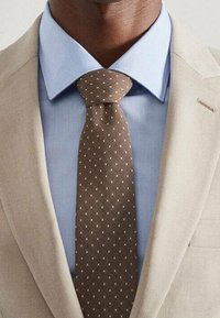 Beige colbert over een lichtblauw shirt, met een bruine stropdas met witte stippen. Netjes zichtbaar is de kraag en de knoop van de stropdas.