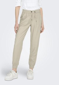 Pantalons de jogging en coton beige avec une taille élastique, cordon de serrage ajustable et poignets côtelés. Associés à des baskets blanches.