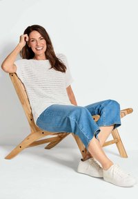 Witte gebreide top met korte mouwen, lichtblauwe cropped jeans en witte sneakers. Model zit op een licht houten stoel met een lattenontwerp.