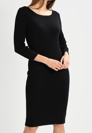 Femme portant une robe en maille noire ajustée tombant au niveau du genou avec manches longues sur un fond clair uni.