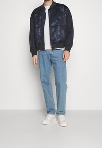 En person står iförd en mörk kamouflage bomberjacka, vit t-shirt, ljusblå jeans och vita sneakers framför en enkel bakgrund.