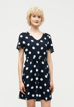 Lyshåret person iført en sort kortærmet kjole med store hvide polkaprikkker og bundet talje, stående foran en ensfarvet baggrund.