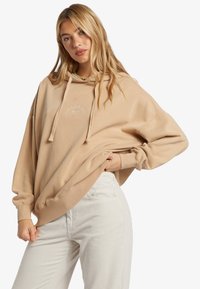 Sudadera beige de corte holgado, con un logo frontal en blanco, cordones y puños de canalé. Combinada con pantalones de pana de color claro.