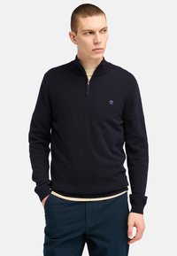 Navy blue zip-neck pullover sweater made of a soft, knitted material. Features a subtle logo on the chest and ribbed cuffs and hem.