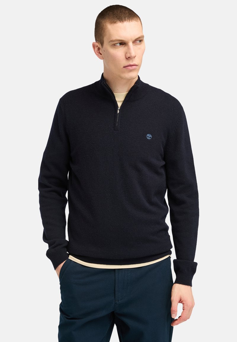 Navy blue zip-neck pullover sweater made of a soft, knitted material. Features a subtle logo on the chest and ribbed cuffs and hem.