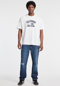 Bílý bavlněný tričko s námořnickou grafikou, která uvádí "CALIFORNIA 81 GUESS JEANS." Nosené s modrými roztrhanými džíny a černými teniskami.