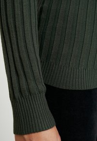 Pull en tricot côtelé vert foncé avec des poignets ajustés, présentant un motif texturé. Accent sur les détails des manches et des poignets sur un fond noir.