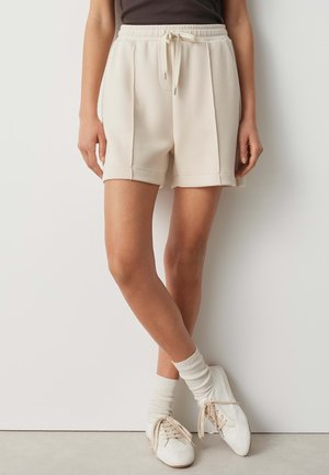 Vrouw draagt beige short met trekkoord, witte sokken en witte sneakers, staat met gekruiste benen tegen een witte muur.