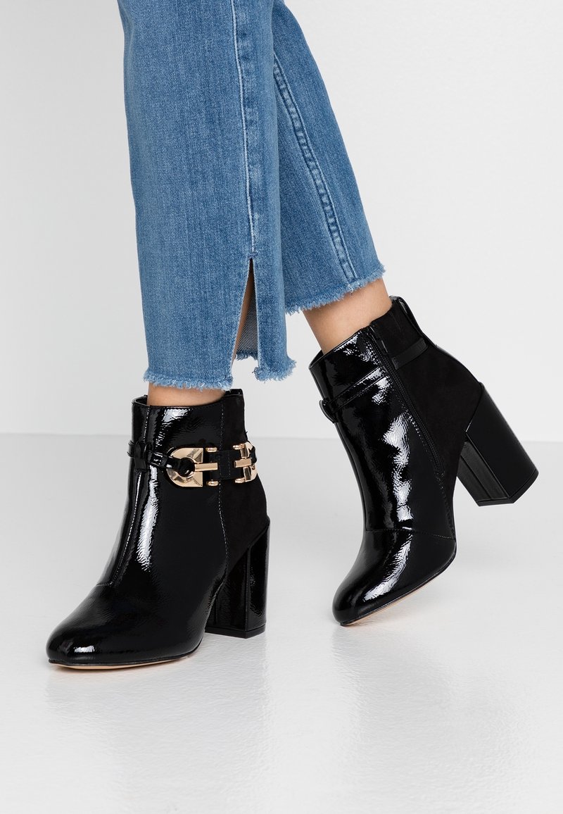 Bottines en cuir verni noir brillant avec détail boucle dorée portées avec un jean denim bleu aux ourlets effilochés et fentes latérales.