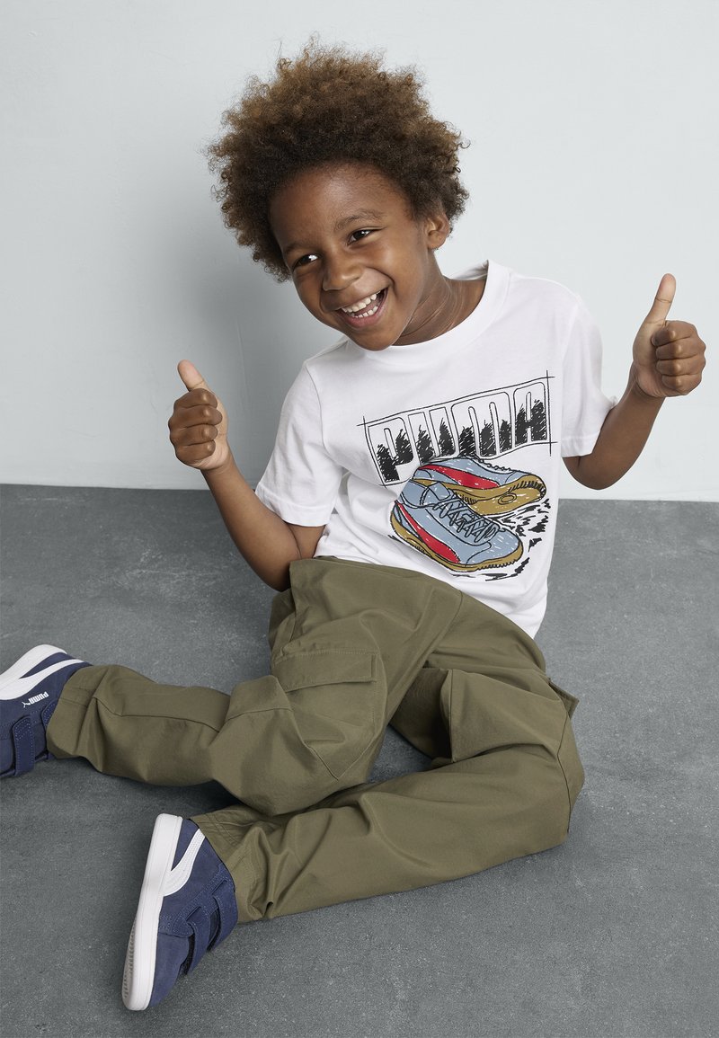 T-shirt blanc avec un graphique de chaussure coloré, pantalon cargo vert olive et baskets navy avec des accents blancs. Assis sur une surface grise.