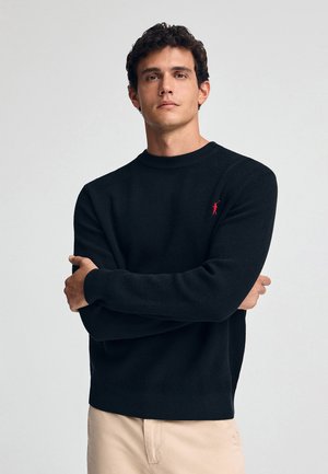 Zwart geribbeld gebreid sweater met een ronde hals, voorzien van een klein rood logo op de borst. Zachte textuur en een ontspannen pasvorm.